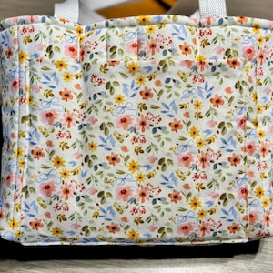 May include: A quilted tote bag with a floral pattern. The bag features a white background with a repeating pattern of pink, yellow, and blue flowers and green leaves. It has white handles and a small pocket on the front.
