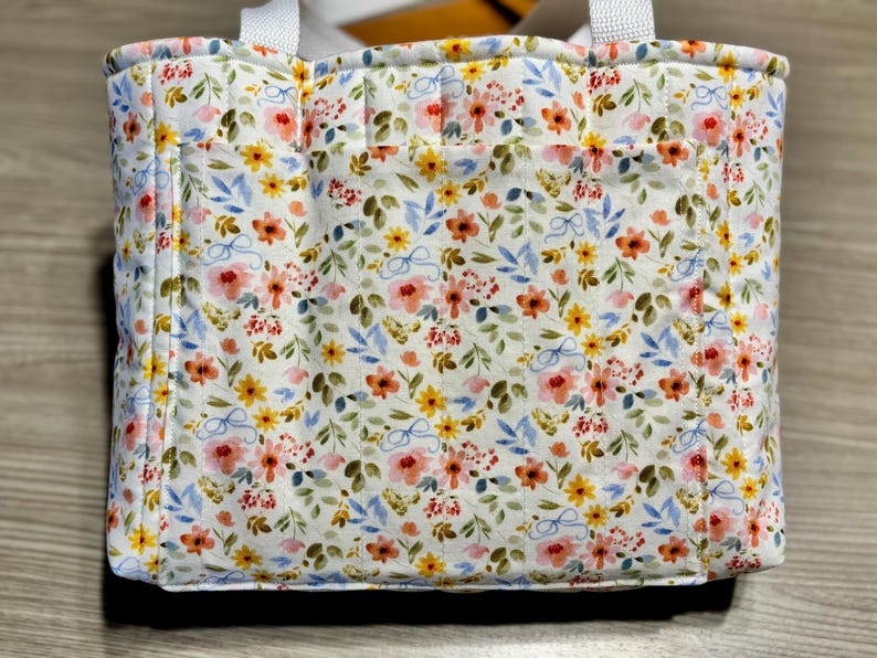 May include: A quilted tote bag with a floral pattern. The bag features a white background with a repeating pattern of pink, yellow, and blue flowers and green leaves. It has white handles and a small pocket on the front.