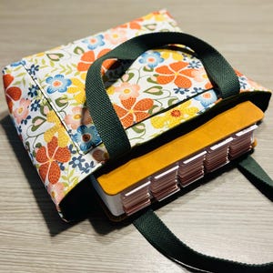 May include: A floral print tote bag with a dark green handle and strap. The bag features a colorful pattern of orange, yellow, and blue flowers on a white background. Inside the bag is a book with labeled tabs.