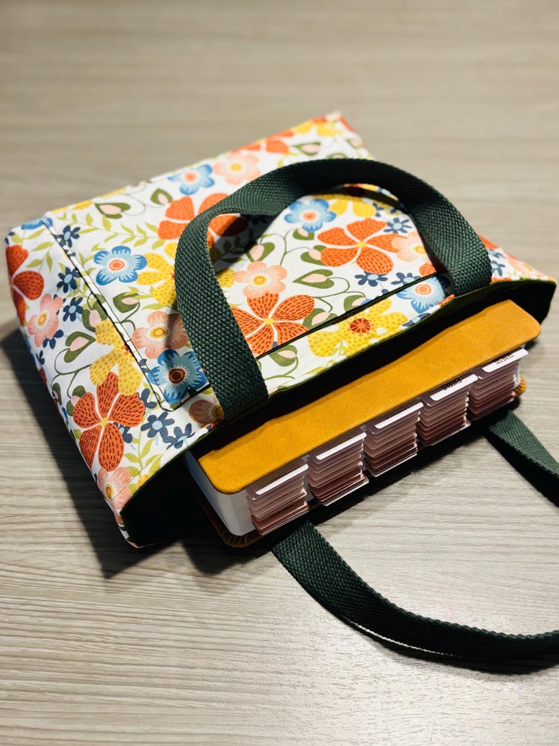 May include: A floral print tote bag with a dark green handle and strap. The bag features a colorful pattern of orange, yellow, and blue flowers on a white background. Inside the bag is a book with labeled tabs.