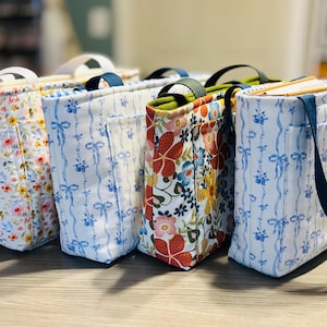 May include: Four fabric tote bags with different floral patterns. One bag has a white background with pink and yellow flowers. Another has a blue and white ribbon pattern. The third has a red, blue, and yellow floral pattern. The fourth has a blue strap.