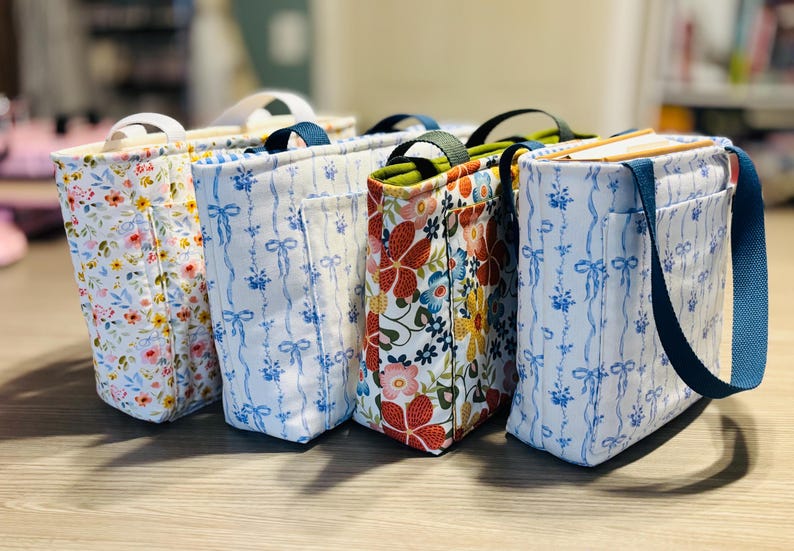 May include: Four fabric tote bags with different floral patterns. One bag has a white background with pink and yellow flowers. Another has a blue and white ribbon pattern. The third has a red, blue, and yellow floral pattern. The fourth has a blue strap.