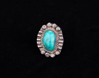 May include: Oval turquoise ring with a silver setting. The turquoise stone is a vibrant teal color, and the silver setting has a detailed design with a rope-like border and small circular accents. The ring is set against a black background.