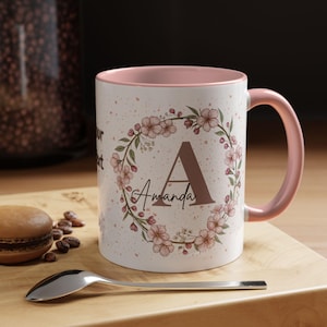 Puede incluir: Taza de cerámica blanca con interior y asa rosas. La taza presenta un diseño de corona floral con la letra "A" y el nombre "Amanda" en marrón. La taza está decorada con detalles florales rosas y pequeñas motas rosas.