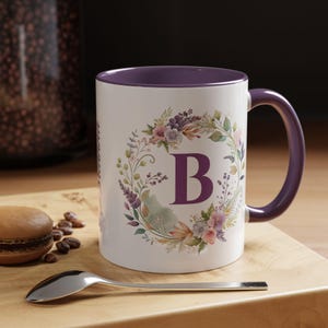 Può includere: Tazza in ceramica bianca con interno e manico viola. La tazza presenta un design a ghirlanda floreale con la lettera "B" al centro. Un cucchiaio e un macaron sono su una superficie di legno.