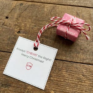 May include: A pink Starburst candy tied with red and white twine, attached to a white tag with the text "Always 'CHEWS' the Right! Merry Christmas!" and a red logo. The items rest on a wooden surface.