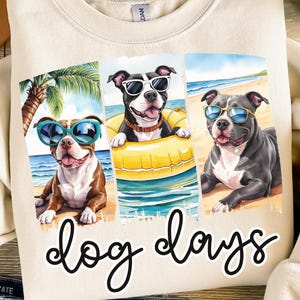 May include: Cream-colored sweatshirt featuring a graphic of three dogs wearing sunglasses on a beach. The dogs are brown and white, black and white, and gray. The words "dog days" are written in a cursive font below the graphic.