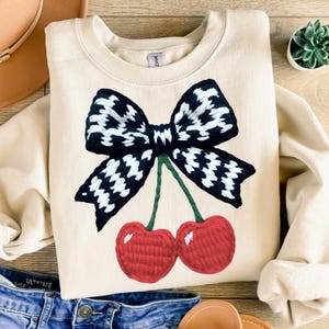 May include: A beige sweatshirt with a black and white checkered bow and two red cherries embroidered on the front.