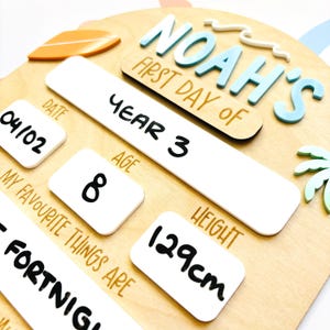 May include: Wooden sign with the text "NOAH'S FIRST DAY OF" in blue and white. Includes sections for date, year, age, and height (129cm). Features a surfboard and palm tree design. The sign reads "Year 3" and "Age 8".