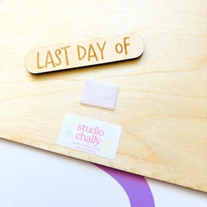 May include: A wooden sign with the words "LAST DAY OF" engraved in a brown font. A small, light pink square of fabric and a white sticker with the text "studio chally" are also present on the light wood surface.