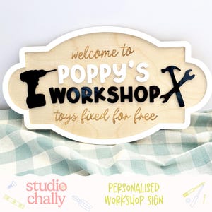 May include: A light-coloured wooden sign with a white border featuring the text "welcome to POPPY'S WORKSHOP toys fixed for free". The sign includes a black silhouette of a power drill on the left and two crossed black wrenches on the right.