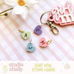 May include: Pastel heart-shaped initial keyring charms. Each charm has a different letter: A, C, J, and M. The charms are attached to a keyring with a lobster clasp. The background is a pink and white striped pattern.
