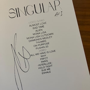 May include: A white sheet of paper with the word "SINGULAR" at the top, followed by a list of song titles. The paper is signed with a black marker. The paper is on a wooden surface.