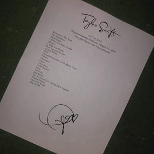 May include: A white sheet of paper with the handwritten signature of Taylor Swift, along with a list of songs from the 1989 World Tour. The date and location, Raymond James Stadium, Tampa, FL, USA, are also printed on the paper.