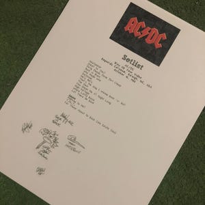Op de afbeelding: Een wit vel papier met het AC/DC-logo en het woord "Setlist". De tekst bevat nummers als "Back in Black" en "Highway to Hell". Er zijn verschillende handtekeningen en tekeningen aanwezig.