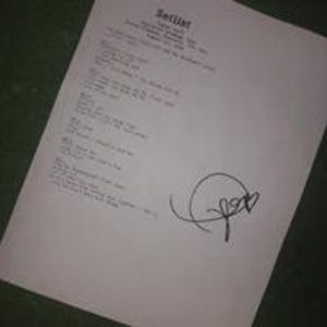 Puede incluir: Una hoja de papel blanca con texto escrito a mano y la palabra "Setlist" en la parte superior. El papel tiene una firma y un pequeño corazón dibujado. El papel está inclinado sobre una superficie oscura.