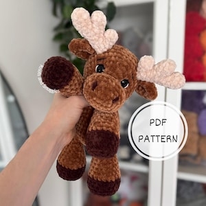 Könnte beinhalten: Ein Elch-Stofftier aus braunem Samtgarn mit hellbeigem Geweih und dunkelbraunen Hufen. Das Spielzeug hat schwarze Sicherheitsaugen und eine gestickte Nase. Der Text auf dem Bild lautet Knots and Snuggles und PDF PATTERN in einem Kreis.