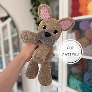Puede incluir: Un juguete de perro de peluche tejido a ganchillo con hilo marrón suave, hocico gris oscuro, interior de orejas rosa y ojos de plástico negro. El juguete tiene detalles de patas y un gráfico circular blanco con el texto PDF PATTERN.