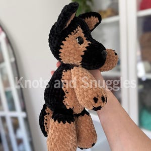 May include: A plush German Shepherd dog toy made of soft, black and tan velvet yarn. The stuffed animal features upright ears, a rounded snout, and dark plastic eyes. The text Knots and Snuggles is overlaid across the center of the image.