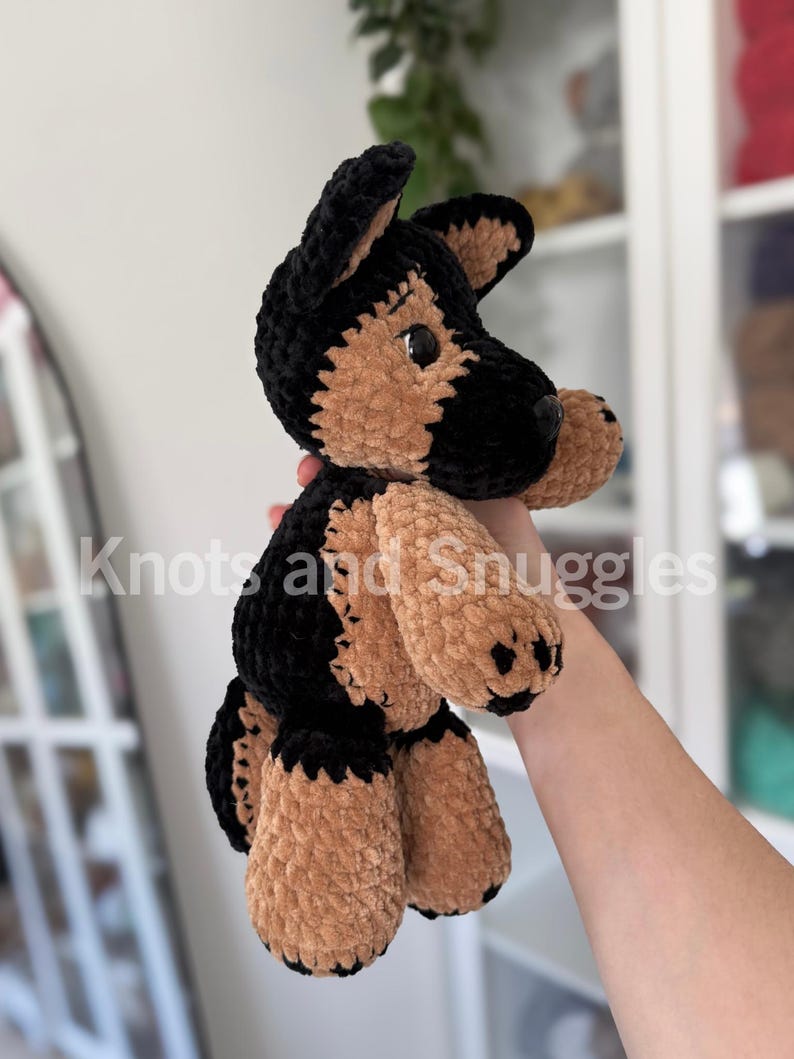 May include: A plush German Shepherd dog toy made of soft, black and tan velvet yarn. The stuffed animal features upright ears, a rounded snout, and dark plastic eyes. The text Knots and Snuggles is overlaid across the center of the image.