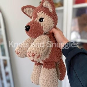May include: A plush crochet dog toy featuring a soft, textured yarn in shades of rust brown and cream. The stuffed animal has pointed ears, a black plastic nose, and black safety eyes. The text Knots and Snuggles is overlaid on the image.