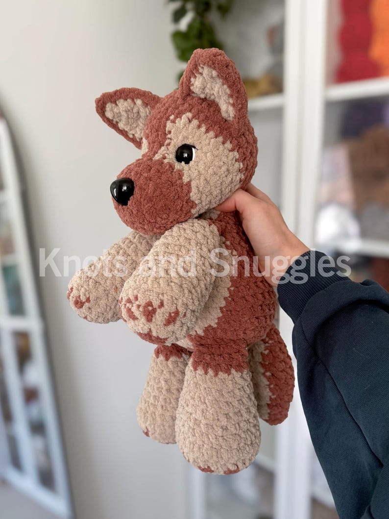 May include: A plush crochet dog toy featuring a soft, textured yarn in shades of rust brown and cream. The stuffed animal has pointed ears, a black plastic nose, and black safety eyes. The text Knots and Snuggles is overlaid on the image.
