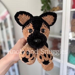 May include: A plush German Shepherd dog toy made of soft, textured yarn in black and tan. The stuffed animal features large, upright ears, black plastic eyes, a black nose, and tan paws with black paw pad details. The text Knots and Snuggles is visible.