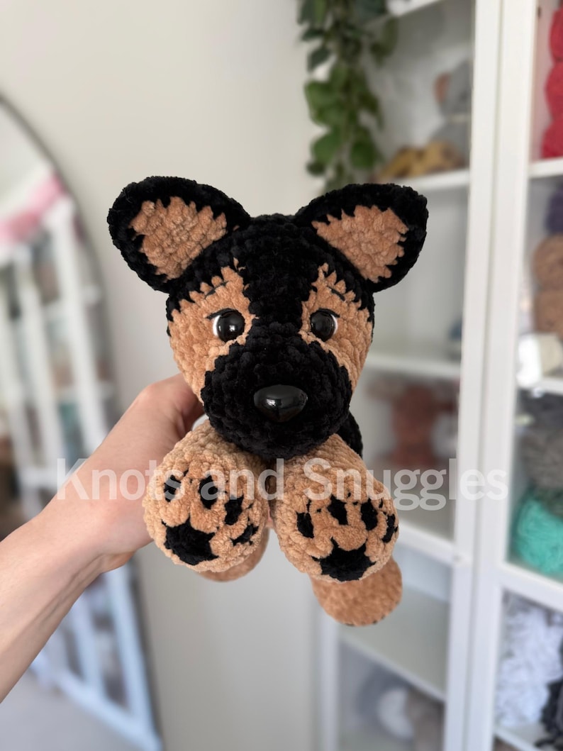 May include: A plush German Shepherd dog toy made of soft, textured yarn in black and tan. The stuffed animal features large, upright ears, black plastic eyes, a black nose, and tan paws with black paw pad details. The text Knots and Snuggles is visible.