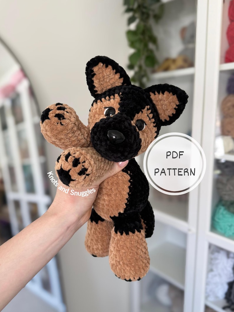 May include: A plush German Shepherd dog toy made of soft brown and black yarn with a black nose and dark eyes. The toy features paw pads with black detailing. A circular graphic on the right side displays the text PDF PATTERN Knots and Snuggles.