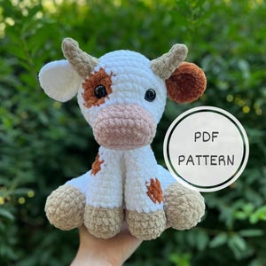 May include: Crocheted stuffed cow toy with white and brown fur. The cow has a pink nose and brown eyes. The toy is being held in a person's hand. PDF PATTERN.