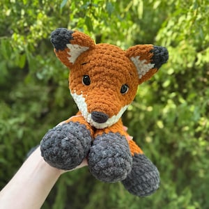Puede incluir: Un peluche de zorro hecho a mano con ganchillo. El zorro tiene pelaje naranja con detalles blancos y negros en las orejas y una nariz negra. Las patas y la parte inferior de las patas son grises. El fondo es follaje verde.