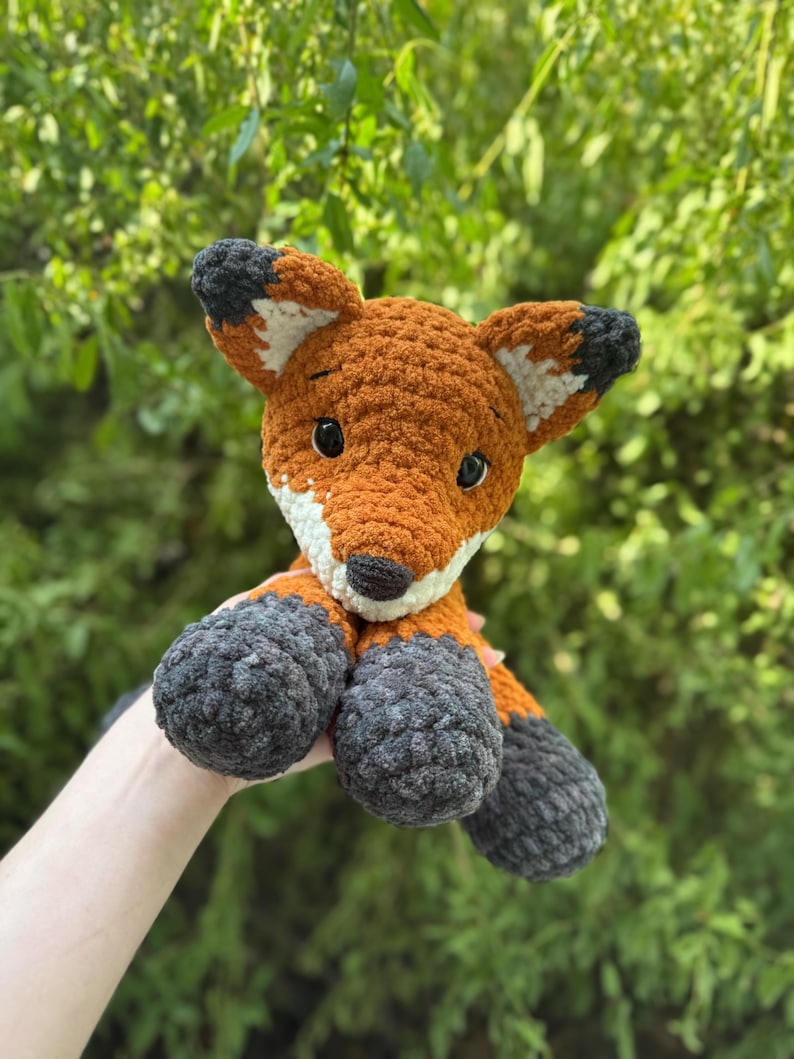 Puede incluir: Un peluche de zorro hecho a mano con ganchillo. El zorro tiene pelaje naranja con detalles blancos y negros en las orejas y una nariz negra. Las patas y la parte inferior de las patas son grises. El fondo es follaje verde.