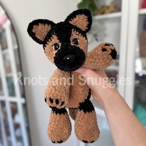 May include: A plush German Shepherd dog toy made of soft, brown and black yarn. The stuffed animal features large, dark plastic eyes, a black nose, and black paw pads. The text Knots and Snuggles is overlaid across the center of the image.