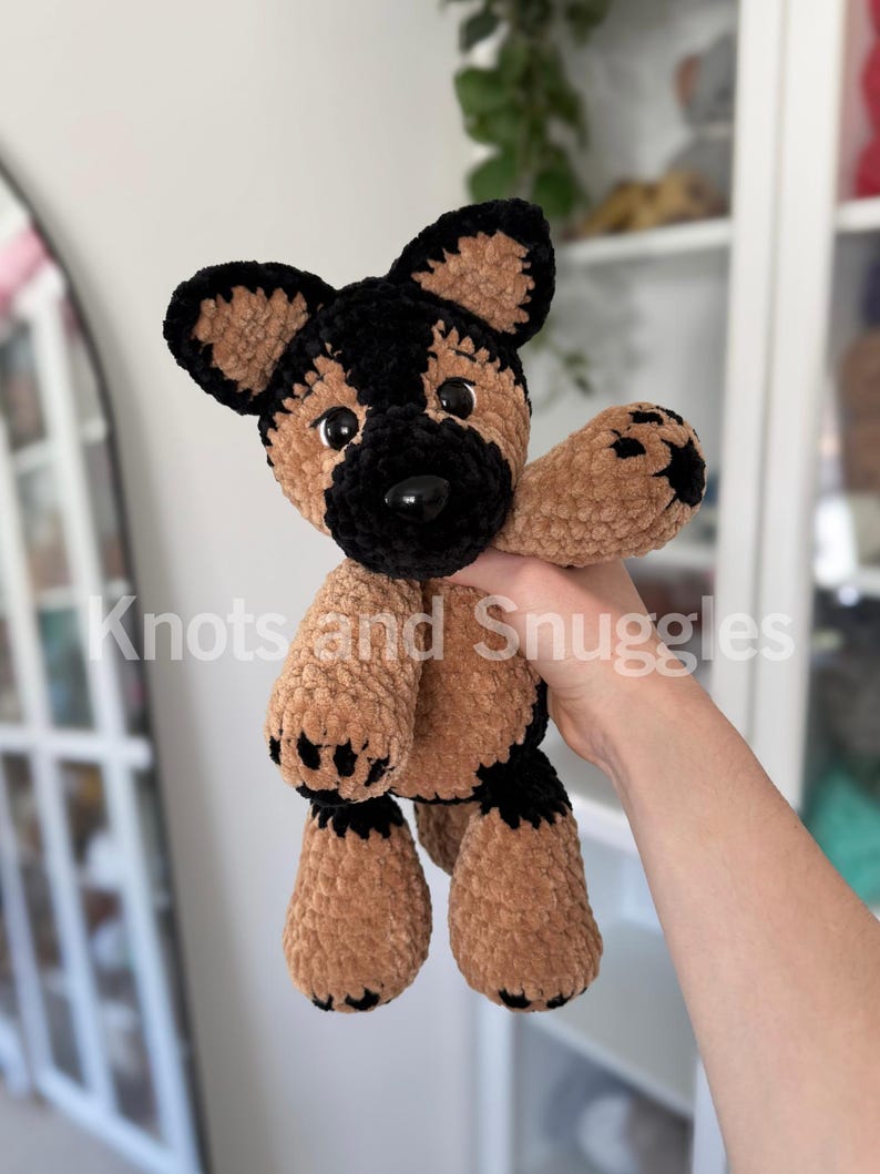 May include: A plush German Shepherd dog toy made of soft, brown and black yarn. The stuffed animal features large, dark plastic eyes, a black nose, and black paw pads. The text Knots and Snuggles is overlaid across the center of the image.