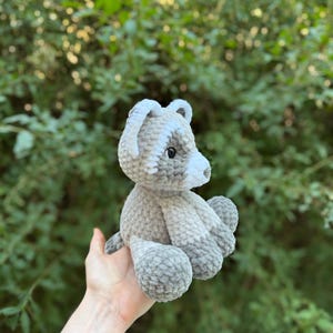 May include: A handmade crochet bear in shades of gray and white. The bear has a rounded body, small ears, and a white snout. The plush toy is held in front of a green leafy background.