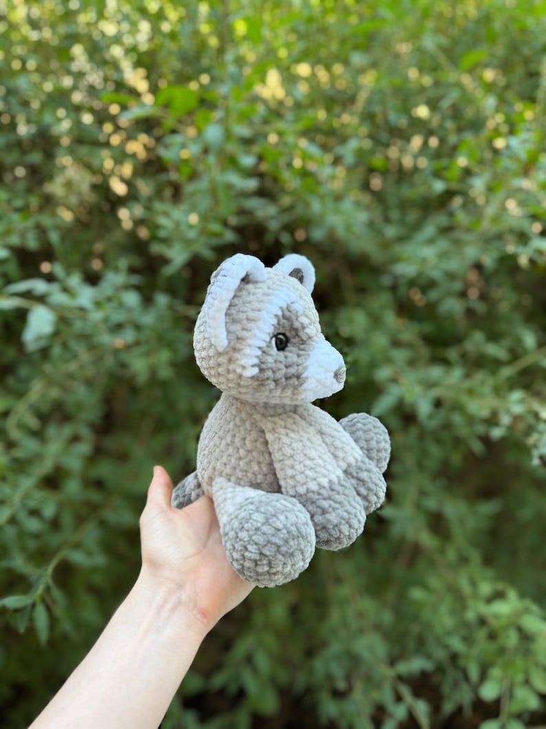 May include: A handmade crochet bear in shades of gray and white. The bear has a rounded body, small ears, and a white snout. The plush toy is held in front of a green leafy background.
