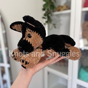 May include: A plush crochet German Shepherd dog toy featuring soft black and tan yarn, dark plastic eyes, and a black nose. The toy is lying down with its front paws extended forward. The text Knots and Snuggles is overlaid on the image.