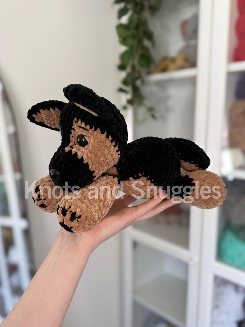 May include: A plush crochet German Shepherd dog toy featuring soft black and tan yarn, dark plastic eyes, and a black nose. The toy is lying down with its front paws extended forward. The text Knots and Snuggles is overlaid on the image.