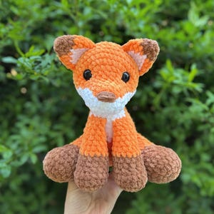 Puede incluir: Un peluche de zorro tejido a mano en naranja y marr&oacute;n con ojos negros, detalles blancos y patas marrones. El zorro est&aacute; sentado erguido con las orejas levantadas. El peluche es un art&iacute;culo lindo y tierno.