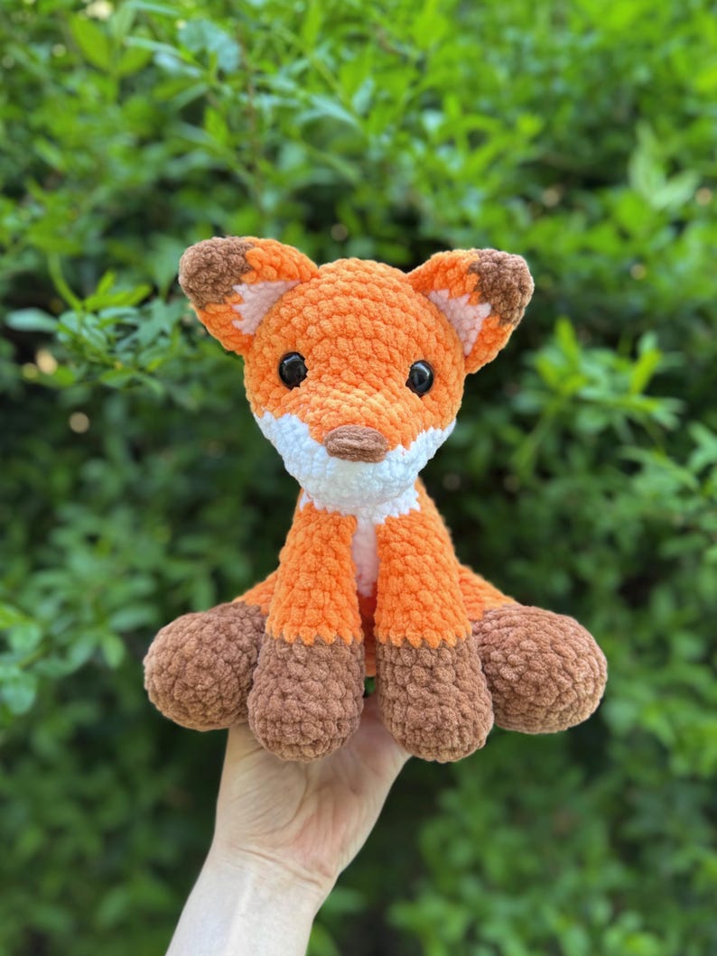 Puede incluir: Un peluche de zorro tejido a mano en naranja y marr&oacute;n con ojos negros, detalles blancos y patas marrones. El zorro est&aacute; sentado erguido con las orejas levantadas. El peluche es un art&iacute;culo lindo y tierno.