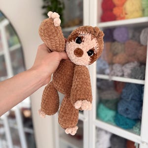 May include: A plush sloth toy crafted from brown and beige chenille yarn. The stuffed animal features a round face with a light beige muzzle, dark brown nose, and large black safety eyes with stitched eyelashes. It has long arms and legs.