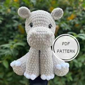 Op de afbeelding: Een gehaakt grijs nijlpaard knuffeldier met witte accenten. Het speelgoed zit op een hand en heeft een witte achtergrond met de tekst "PDF PATTERN".