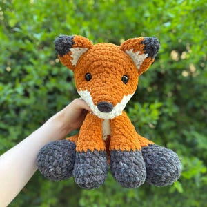 Puede incluir: Un zorro de crochet hecho a mano, con pelo naranja, blanco y gris. El zorro tiene ojos negros y un hocico blanco. Las orejas y los pies tienen detalles grises y blancos. El fondo es follaje verde.