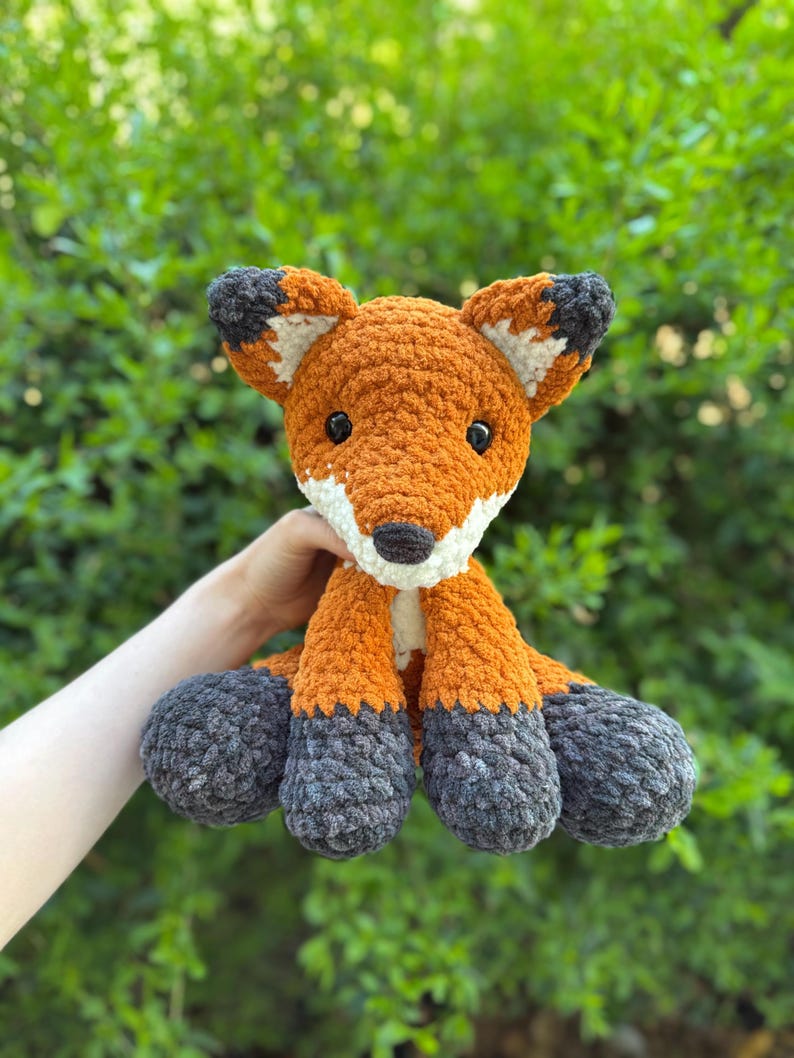 Puede incluir: Un zorro de crochet hecho a mano, con pelo naranja, blanco y gris. El zorro tiene ojos negros y un hocico blanco. Las orejas y los pies tienen detalles grises y blancos. El fondo es follaje verde.
