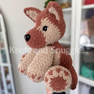 May include: A plush crochet dog toy featuring a two-tone design of dark brown and light beige yarn. The stuffed animal has pointed ears, a black plastic nose, and black safety eyes. The text Knots and Snuggles is visible across the center.