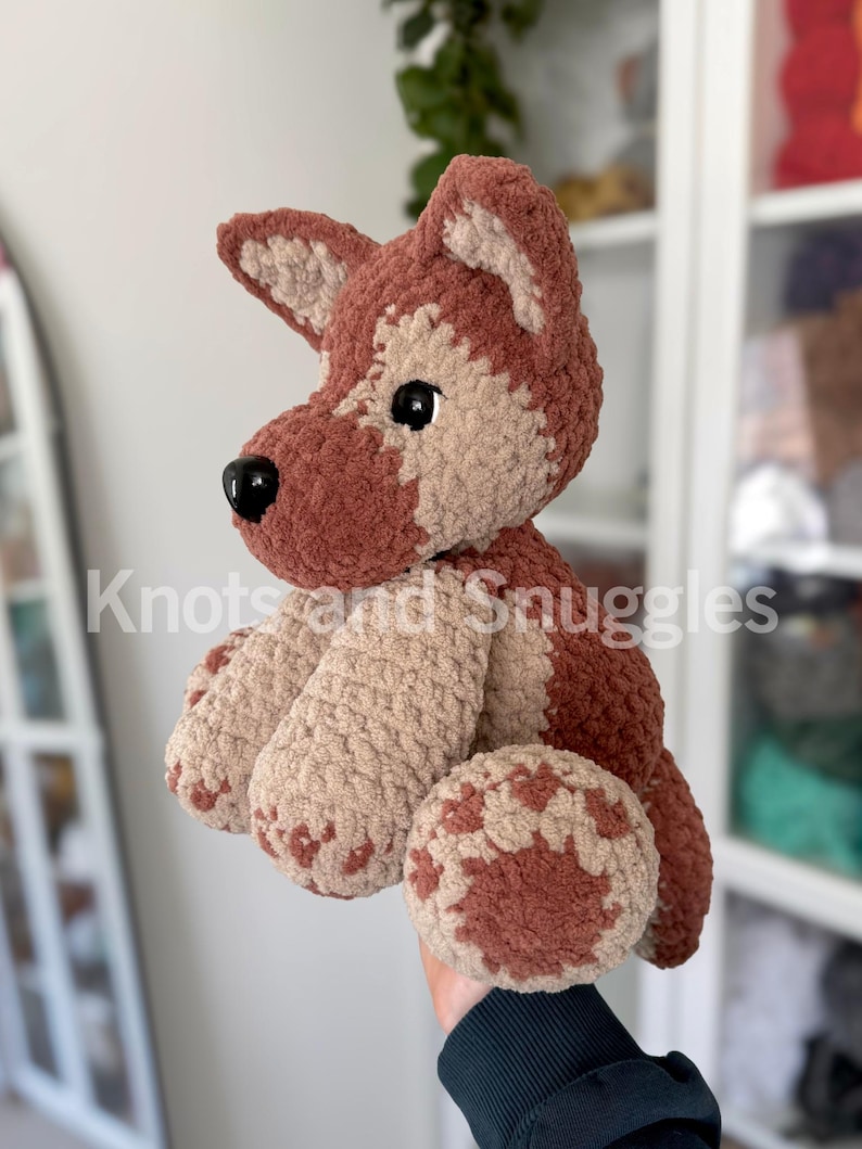 May include: A plush crochet dog toy featuring a two-tone design of dark brown and light beige yarn. The stuffed animal has pointed ears, a black plastic nose, and black safety eyes. The text Knots and Snuggles is visible across the center.