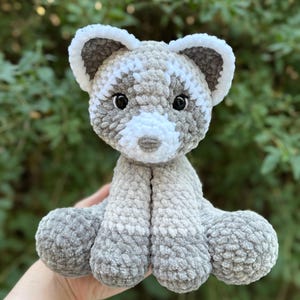 May include: A handmade crochet raccoon plush toy in shades of gray and white. The plush has a soft, textured appearance with black safety eyes and a white muzzle. The raccoon is sitting with its arms and legs outstretched.