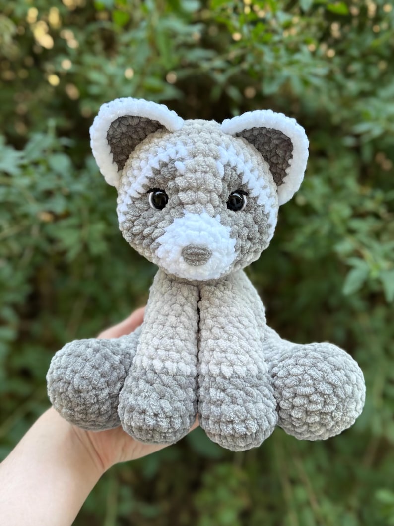 May include: A handmade crochet raccoon plush toy in shades of gray and white. The plush has a soft, textured appearance with black safety eyes and a white muzzle. The raccoon is sitting with its arms and legs outstretched.