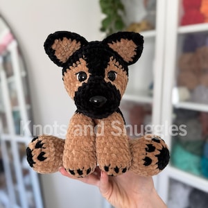 May include: A crocheted German Shepherd plush toy made of soft, tan and black velvet yarn. The stuffed animal features large, dark plastic eyes, a black nose, and black paw pads. The text Knots and Snuggles is visible across the center.