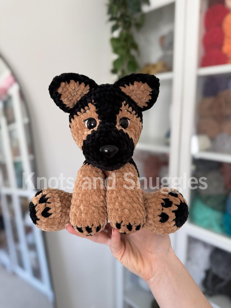 May include: A crocheted German Shepherd plush toy made of soft, tan and black velvet yarn. The stuffed animal features large, dark plastic eyes, a black nose, and black paw pads. The text Knots and Snuggles is visible across the center.